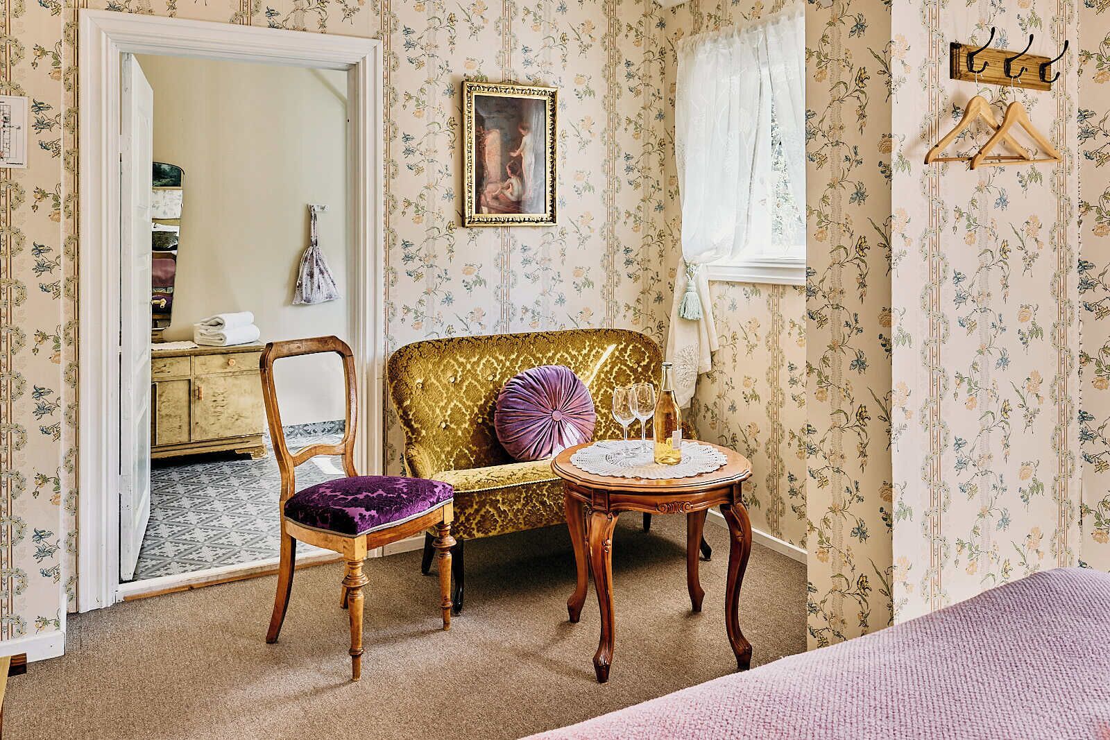 Et værelse med en vintage sofa, en træstol, et lille rundt bord med en flaske og glas samt et spejl på væggen.