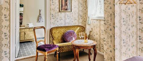 A room with a vintage sofa, a wooden chair, a small round table with a bottle and glasses, and a mirror on the wall.
