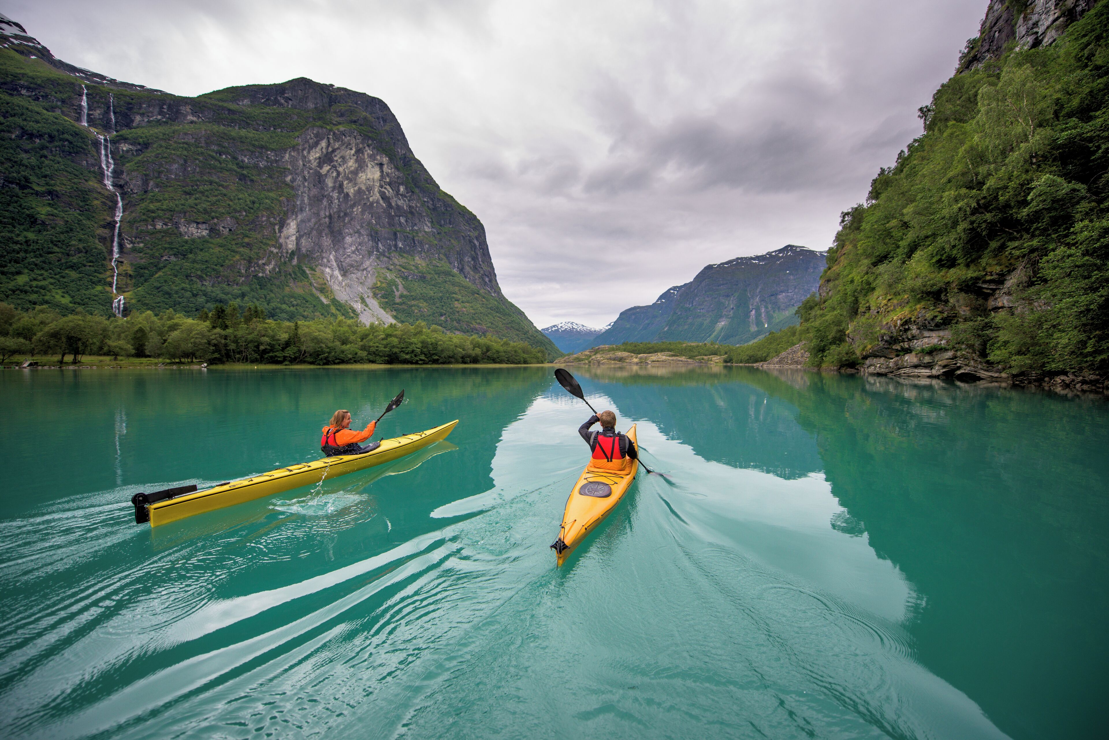 Two kayakers on a calm river with mountainous scenery in the background.