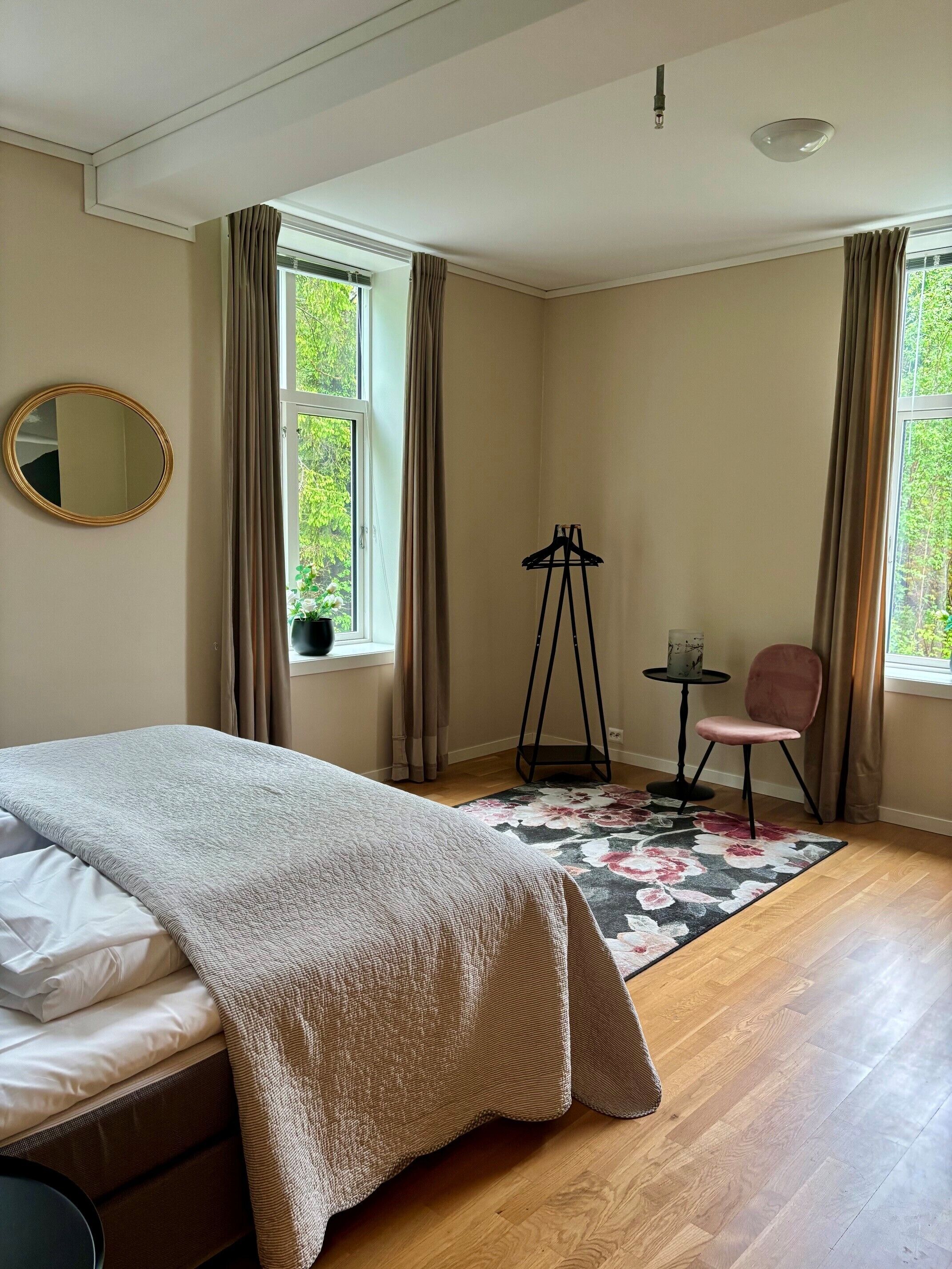 A bedroom with a bed, a chair, a small table, a mirror, and a window with curtains.