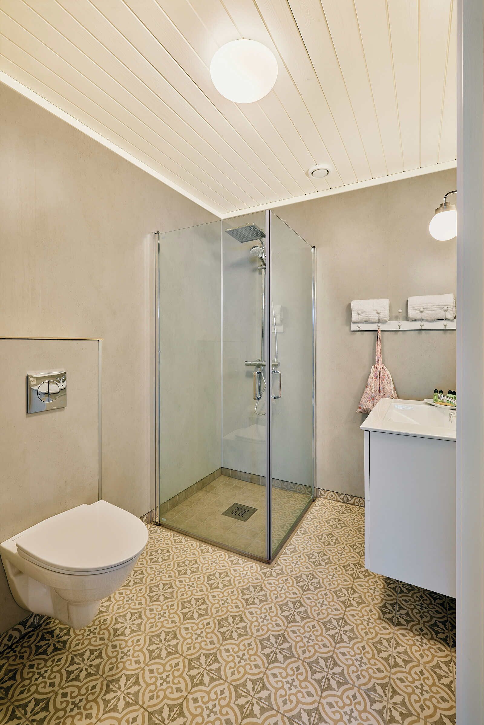 A bathroom with a toilet, a glass shower enclosure, patterned floor tiles, and a sink with a towel rack.