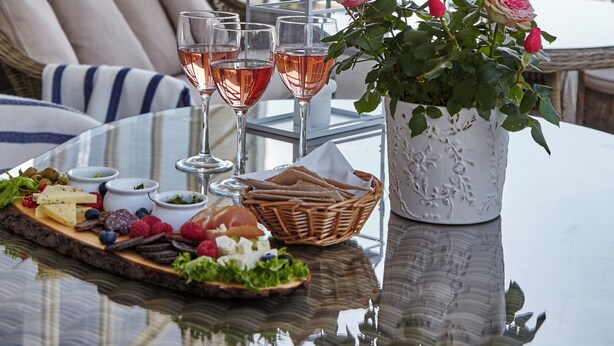 Een gedekte tafel met een kaasplank, fruit en glazen rosé, met uitzicht op een meer en bergen op de achtergrond.