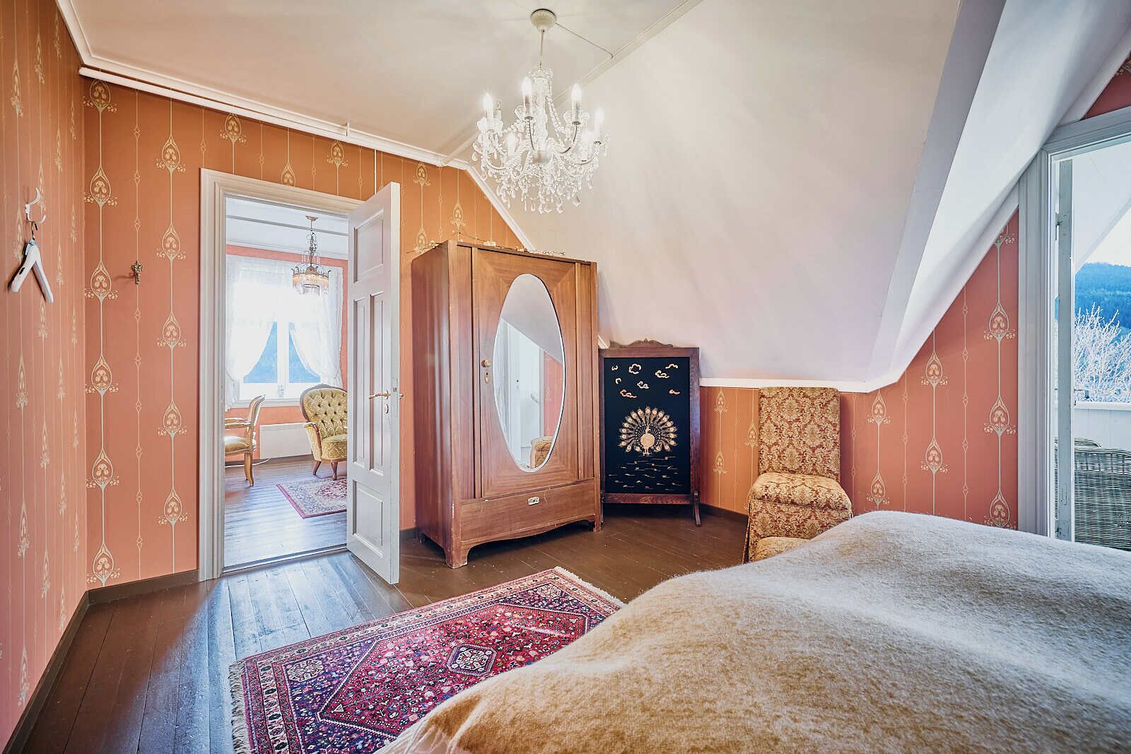 A bedroom with a large bed, a wardrobe, a chair, and a view of the outdoors.