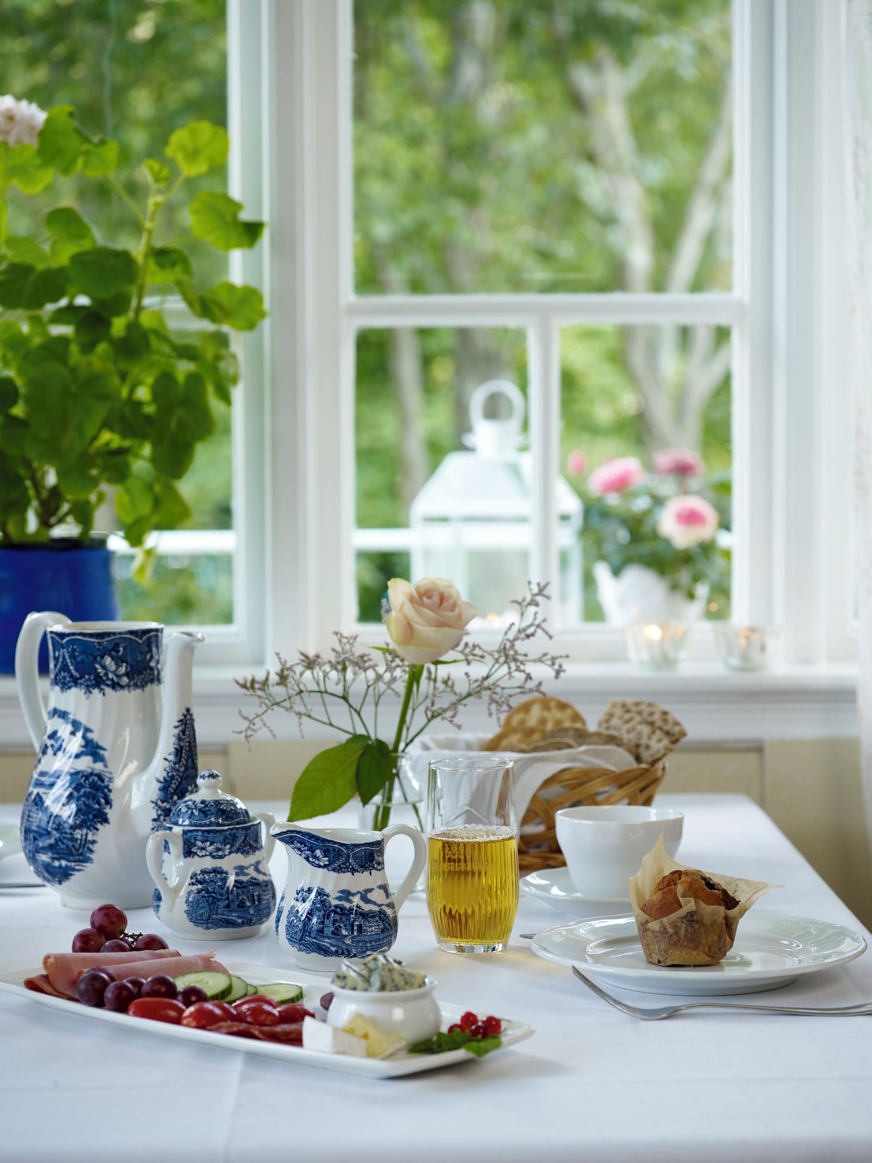 A table set with a variety of food items and drinkware, including a pitcher, teapot, and a glass of a yellow beverage, with a view of greenery outside a window.