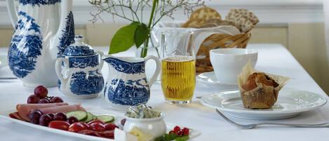 A table set with a variety of food items and drinkware, including a pitcher, teapot, and a glass of a yellow beverage, with a view of greenery outside a window.