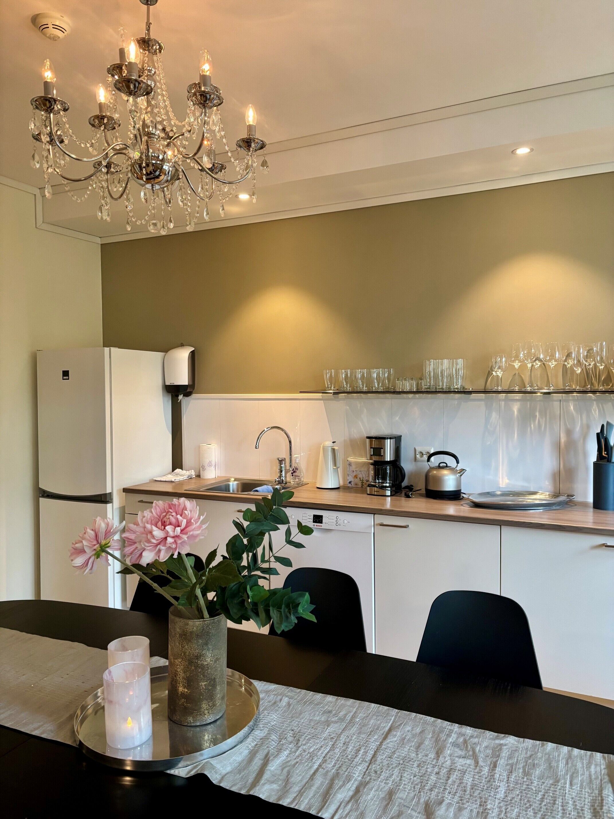 A modern kitchen with a chandelier, white cabinets, a refrigerator, a sink, and a dining area with a table set for two.