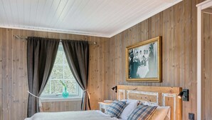 Een slaapkamer met houten lambrisering, een bed, een raam met gordijnen, een nachtkastje en een wandlamp.