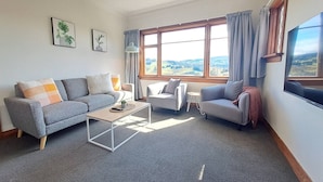 A living room with a grey sofa, a small table, two armchairs, and a large window with curtains. - Spacious, Affordable, Peaceful Family Home (Dunedin)