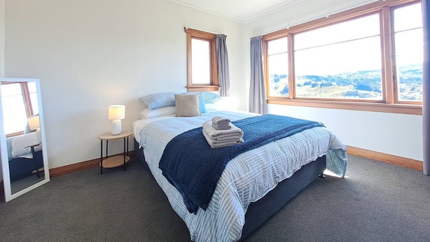 A bedroom with a large bed, a bedside table with a lamp, a mirror, and two windows with curtains. - Spacious, Affordable, Peaceful Family Home (Dunedin)