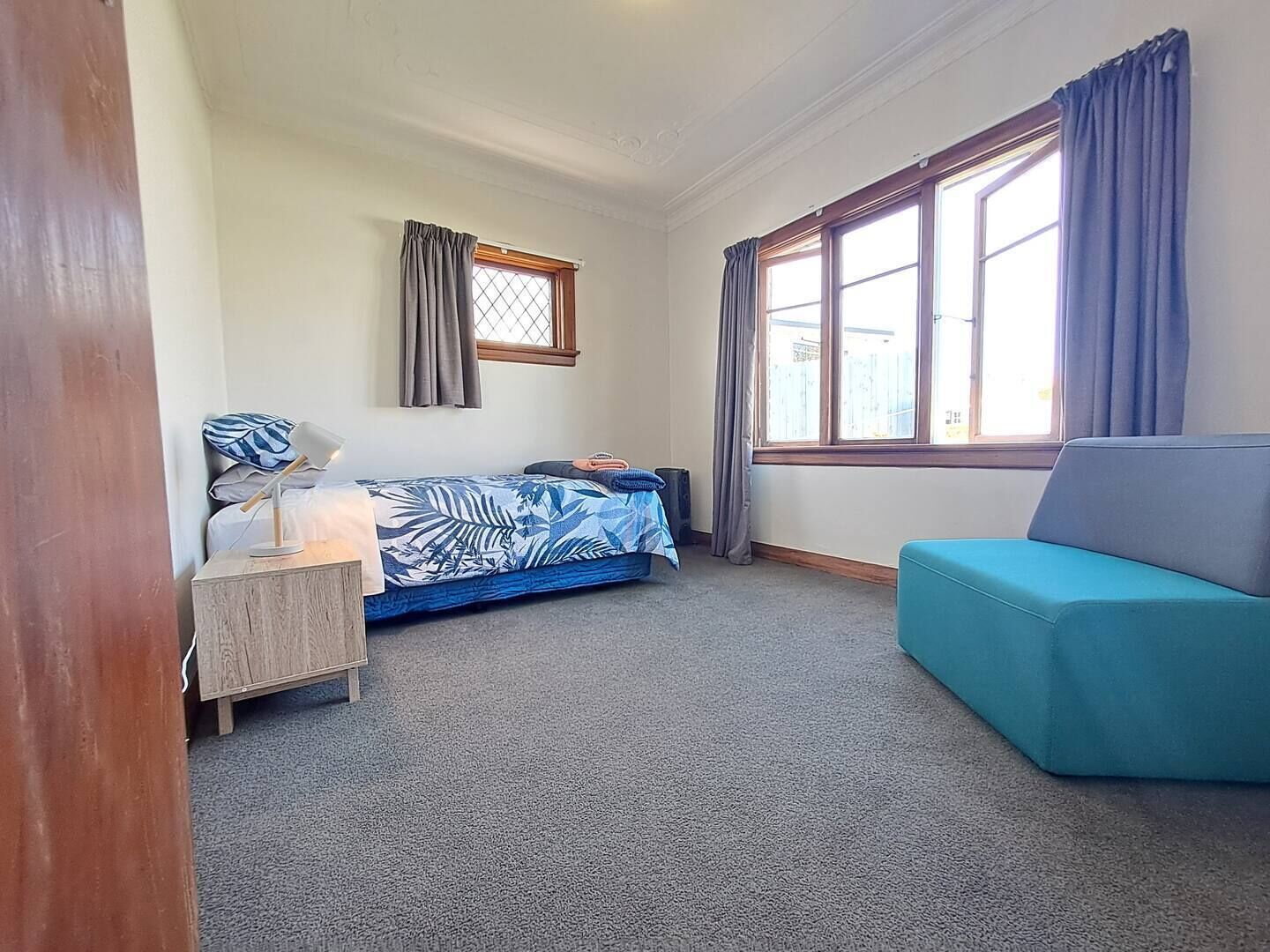 Et lille, godt oplyst værelse med en enkelt seng, et skrivebord med lampe, en blå sofa og store vinduer med gardiner.