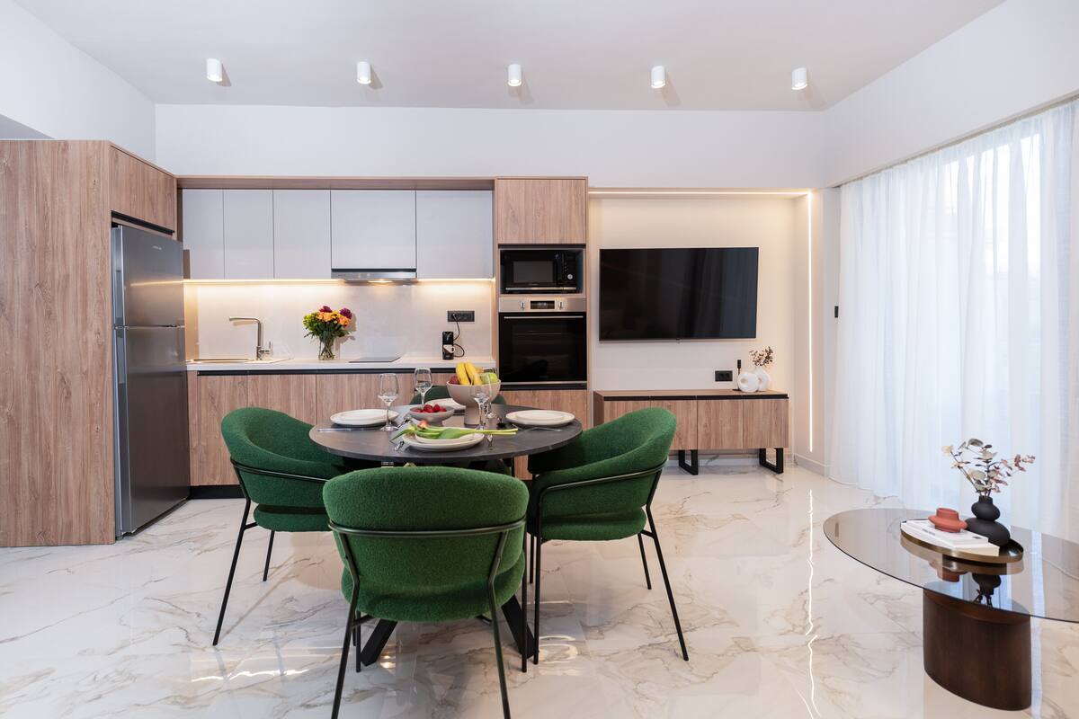 A modern kitchen with a dining area, featuring a marble floor, green chairs, and a glass table.