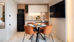 Une cuisine moderne avec un coin repas, comprenant un réfrigérateur, un four à micro-ondes et un four de couleur noire, une table ronde avec des chaises orange et un téléviseur à écran plat fixé au mur.