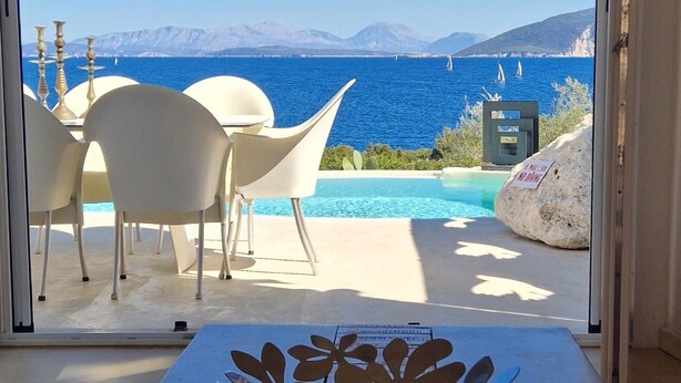 Una stanza con vista sul mare, una piscina e un'area pranzo con sedie e tavolo bianchi.