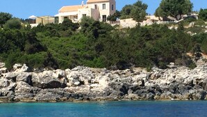 Una proprietà costiera con una grande casa su un pendio roccioso, affacciata su un mare cristallino e blu.