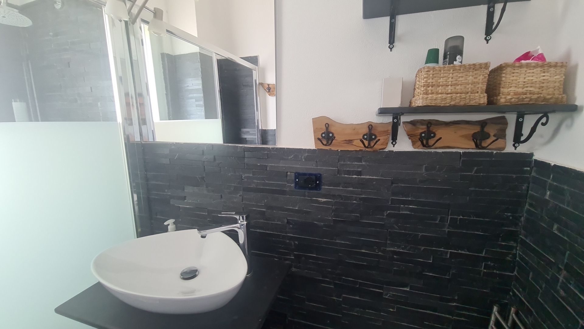 A bathroom with a white sink, black tiled walls, and a wooden shelf with toiletries.