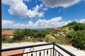 Un balcón con vistas a un paisaje rural, que incluye casas, árboles y un cielo azul despejado.