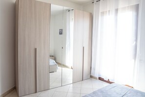Ein Schlafzimmer mit großem Kleiderschrank, einem Bett und einem Fenster mit durchsichtigen Vorhängen.