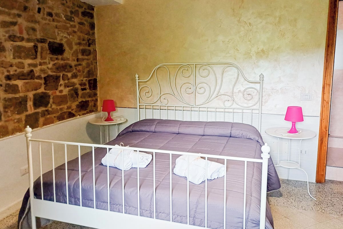 Un dormitorio con pared de piedra, estructura de cama de metal blanco, colcha morada y dos mesitas de noche con lámparas rosadas.