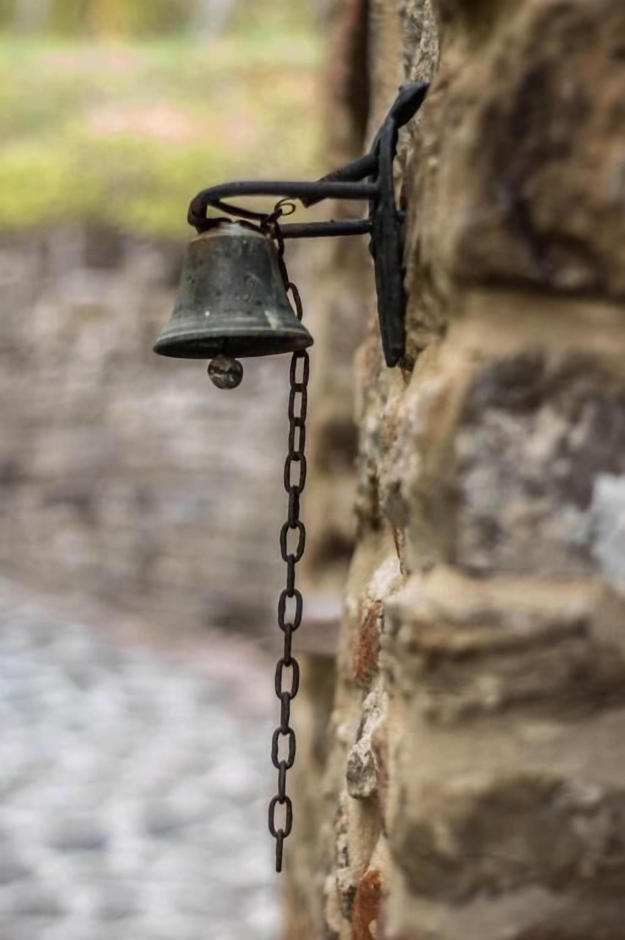 A metal bell hanging from a chain attached to a stone wall.