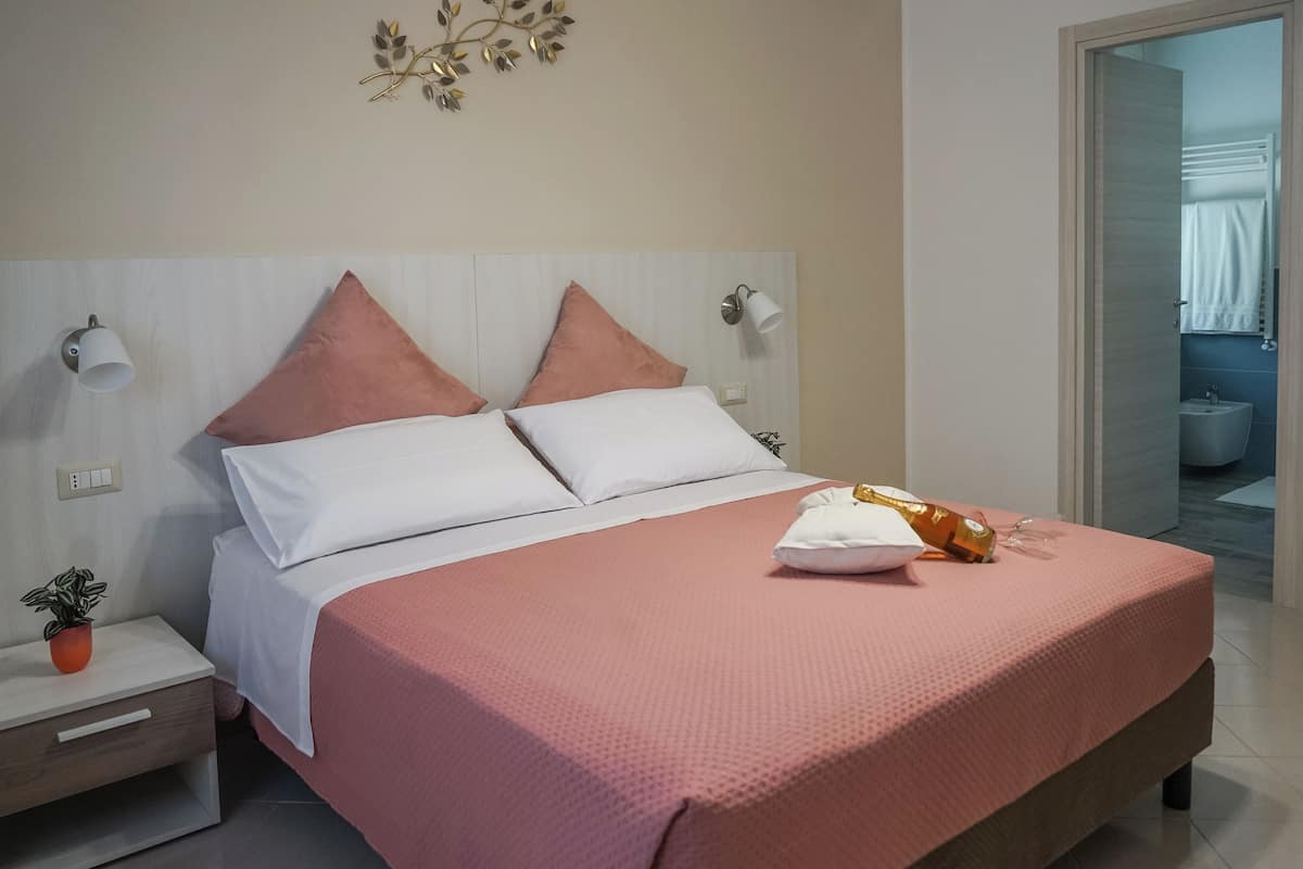 Una cama bien hecha con una colcha rosa y almohadas blancas, una mesita de noche con una planta y una lámpara fijada en la pared.