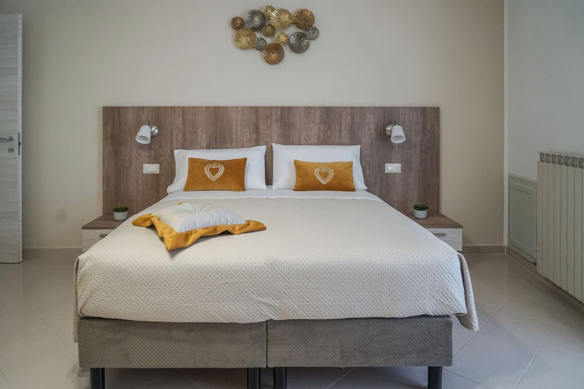 Una cama bien hecha con una decoración en blanco y dorado, dos mesitas de noche con lámparas y un adorno en la pared sobre la cama.