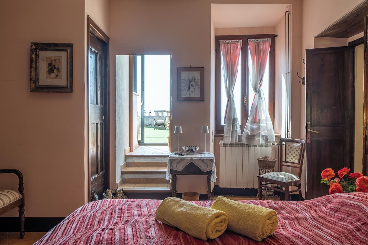 Ein Schlafzimmer mit einem Bett, zwei aufgerollten Handtüchern, einem Stuhl, einem Tisch mit einer Schüssel und einem Fenster mit Vorhängen.