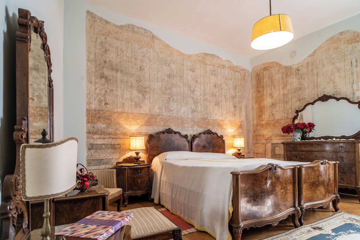 Ein traditionell eingerichtetes Schlafzimmer mit einem großen Bett, einem Holzkopfende und einem Spiegel. Zwei Nachttische mit Lampen und eine Kommode mit Spiegel gehören zur Einrichtung.