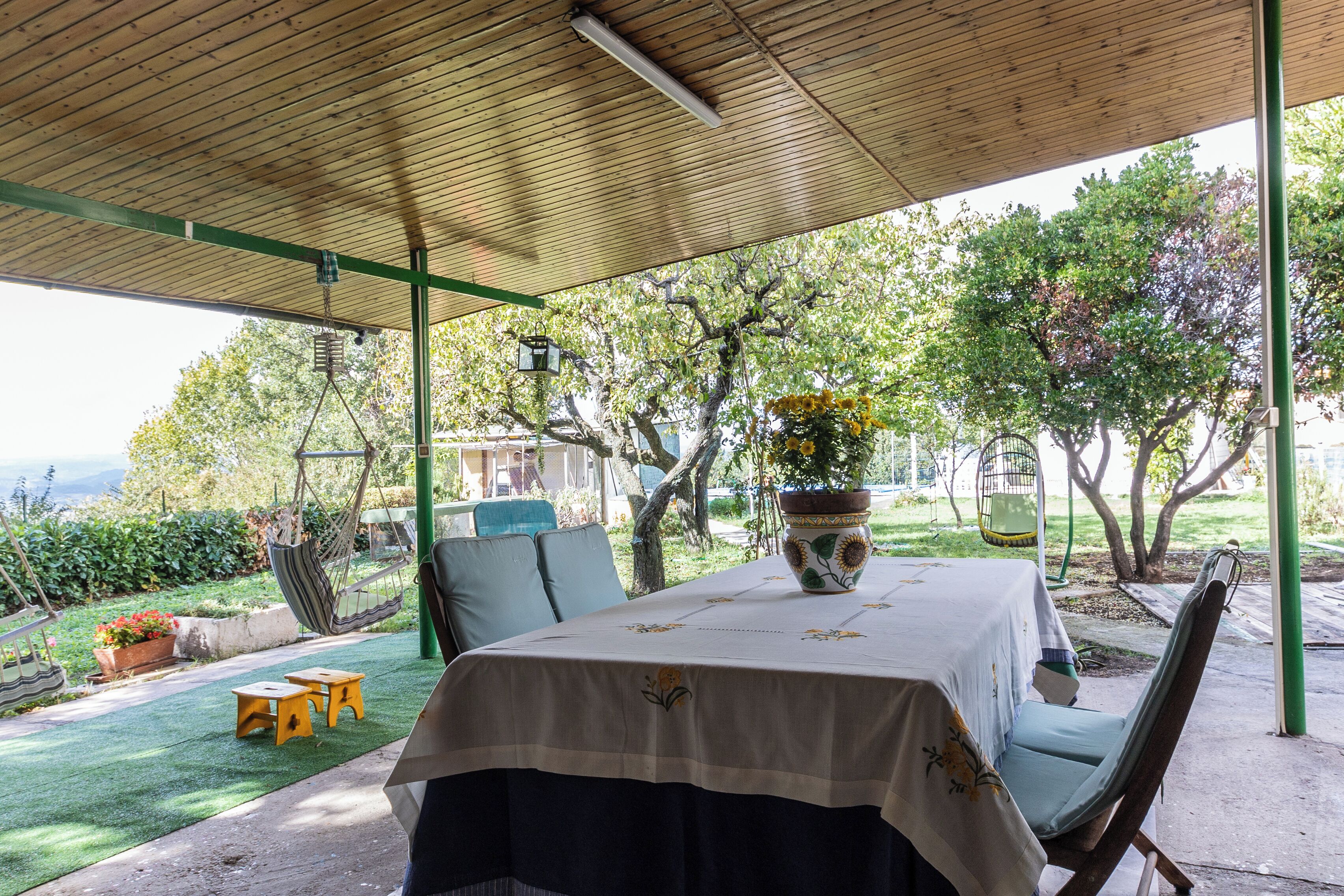 Une terrasse extérieure couverte, avec une table dressée pour le repas, entourée de chaises et agrémentée d’un fauteuil suspendu.