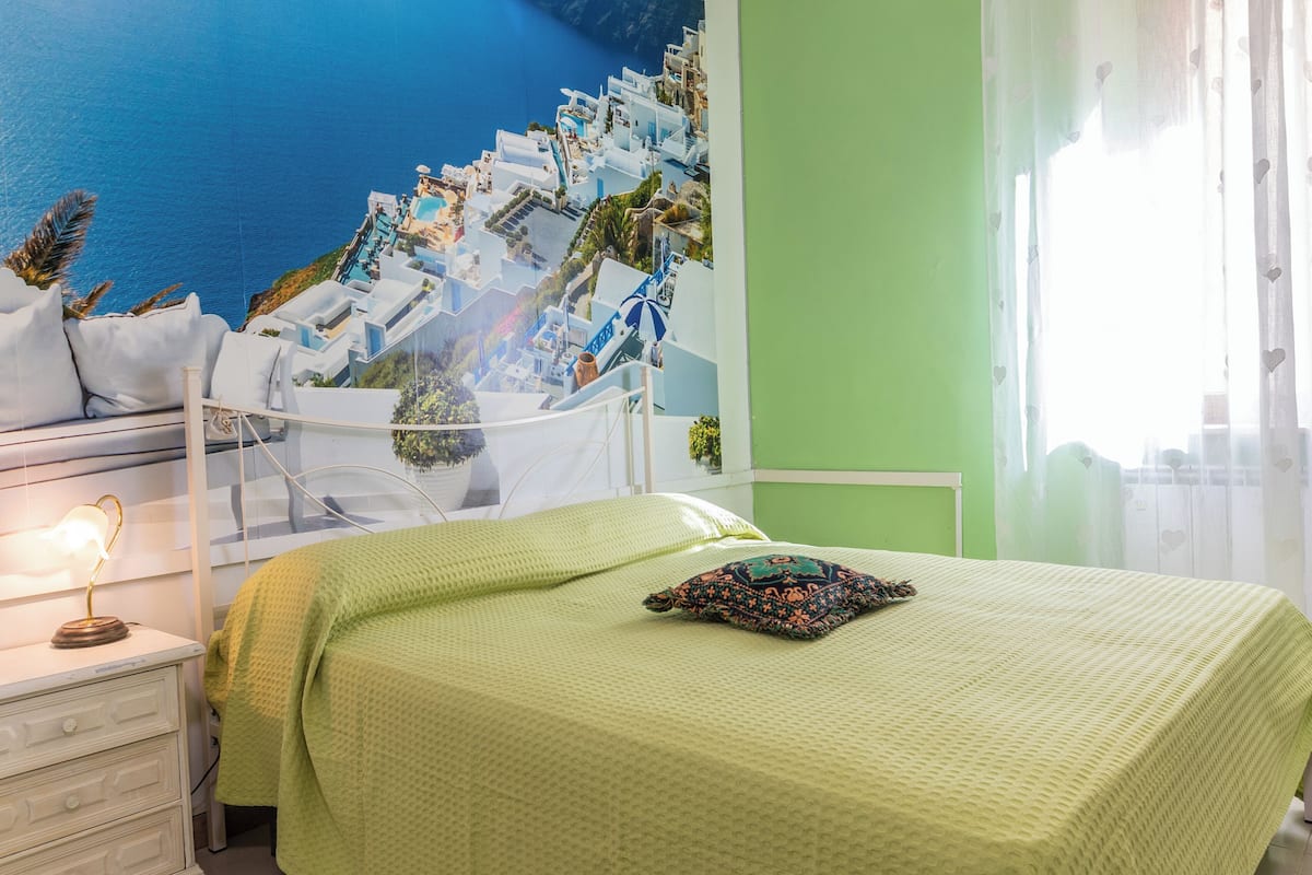 A bedroom with a bed, a nightstand, a lamp, a window with sheer curtains, and a view of a coastal scene.