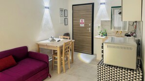 Quarto compacto com sofá roxo, pequena mesa de jantar, porta de madeira com placa de 'Não Fumar', cozinha compacta com pia e um espelho acima.