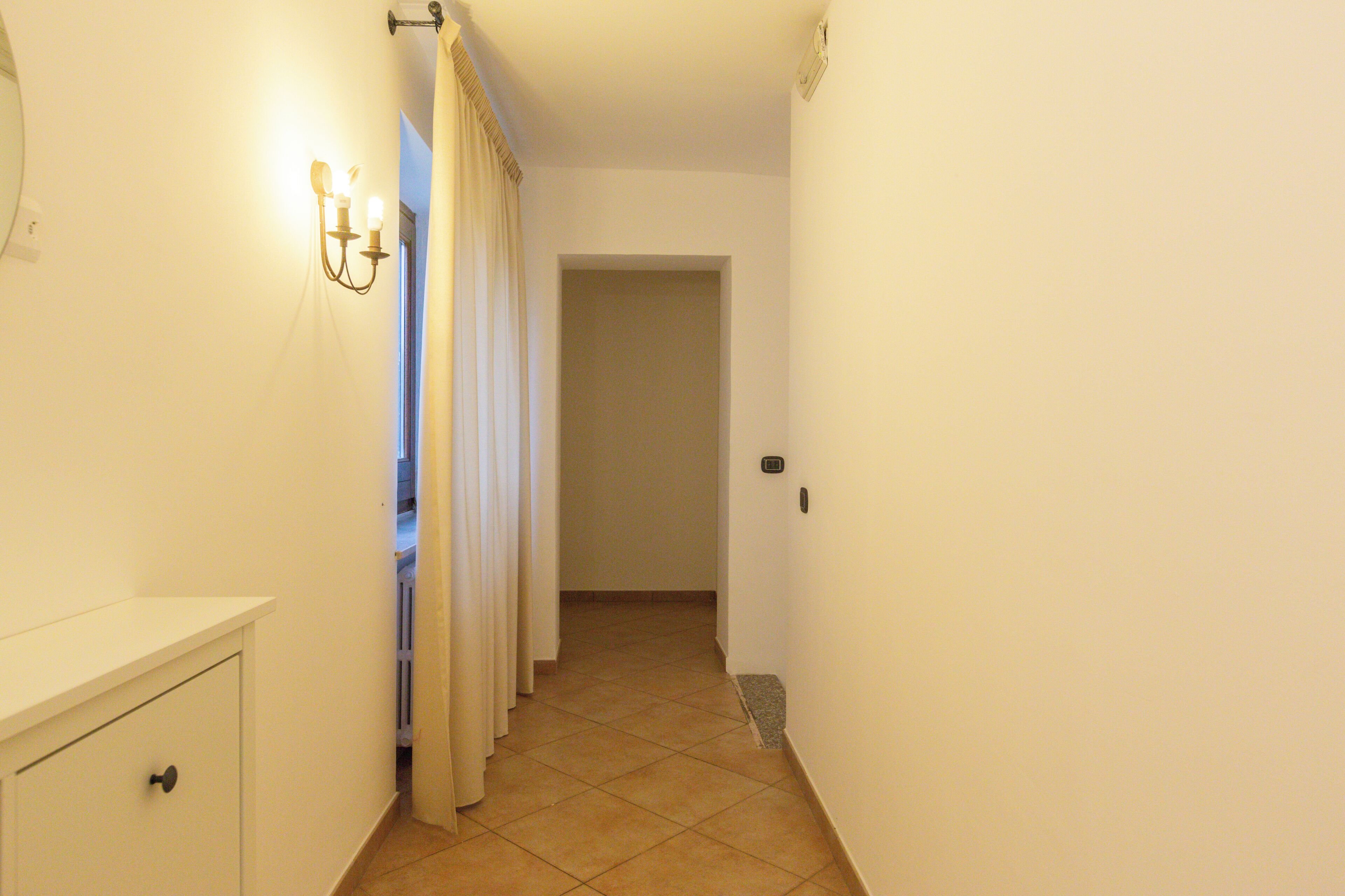 Un couloir étroit, avec une porte blanche au fond, une petite armoire sur la gauche et un luminaire fixé au mur.