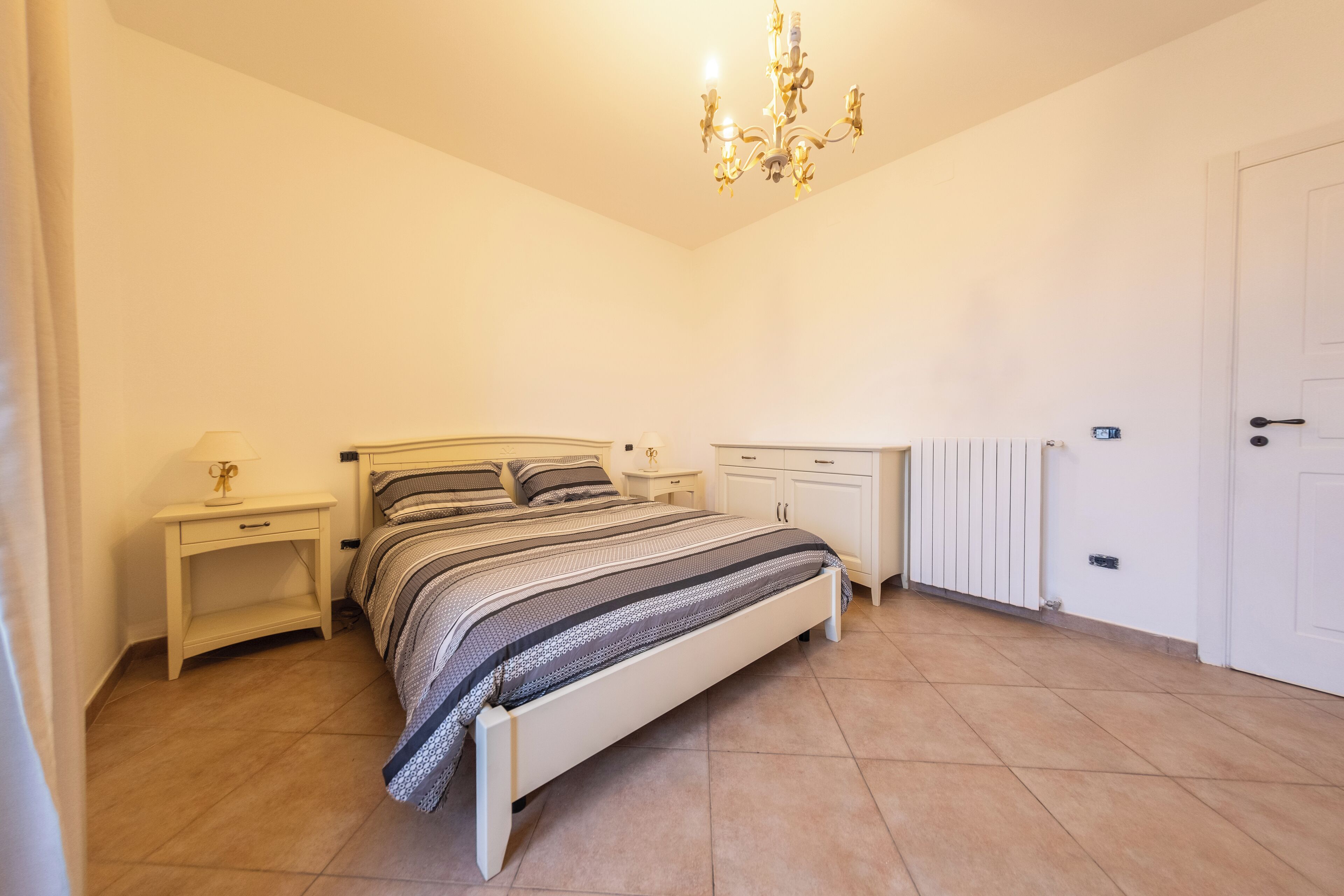 A bedroom with a bed, nightstands, a dresser, a radiator, and a chandelier.