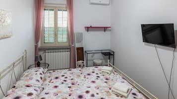 A bedroom with a bed, a window with curtains, a radiator, a TV, and a shelf.