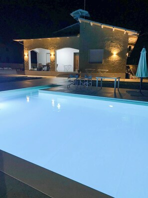 Une piscine avec un espace patio, des sièges extérieurs et un bâtiment doté d’une façade en pierre et d’un éclairage chaleureux.