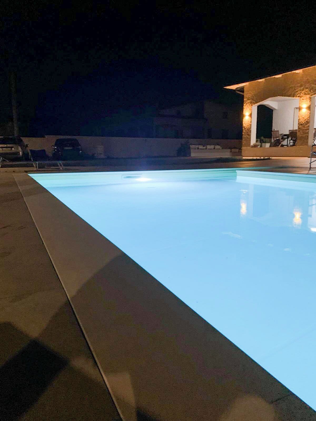 Ein Schwimmbecken bei Nacht mit beleuchtetem Rand und einem überdachten Bereich mit Sitzgelegenheiten.