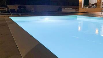 Ein Schwimmbecken bei Nacht mit beleuchtetem Rand und einem überdachten Bereich mit Sitzgelegenheiten.