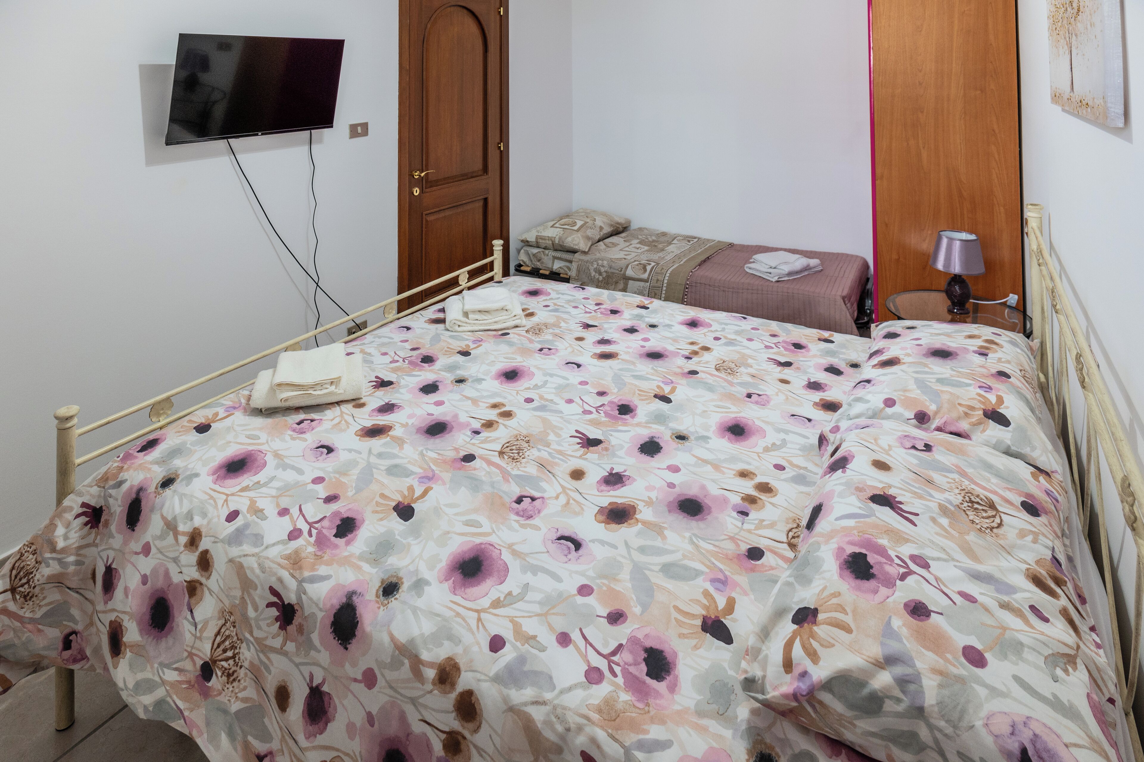 A bedroom with a bed, a TV mounted on the wall, a bedside table with a lamp, and a door.
