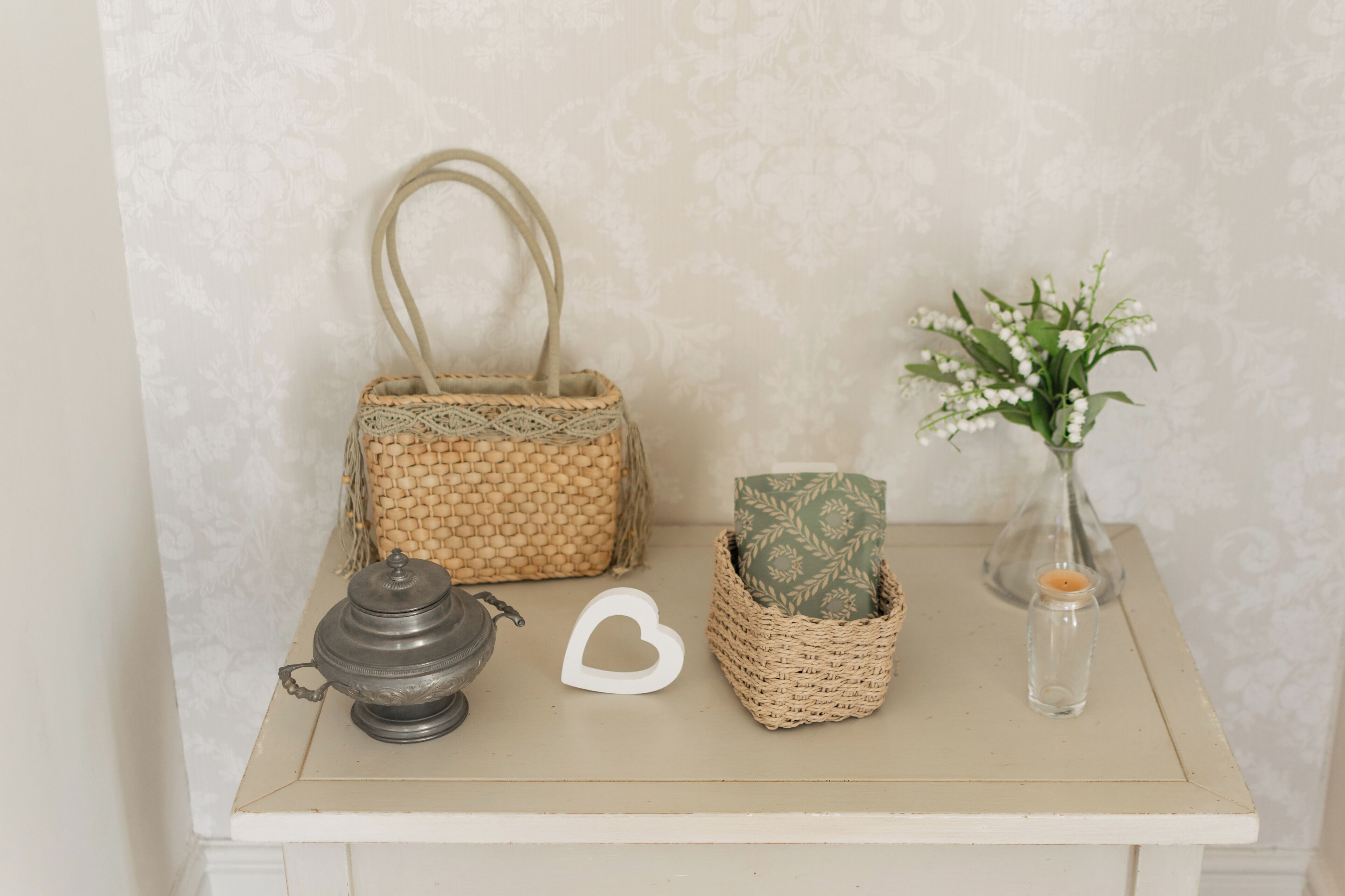 A small table with a woven basket, a candle, a decorative heart, and a vase with flowers.