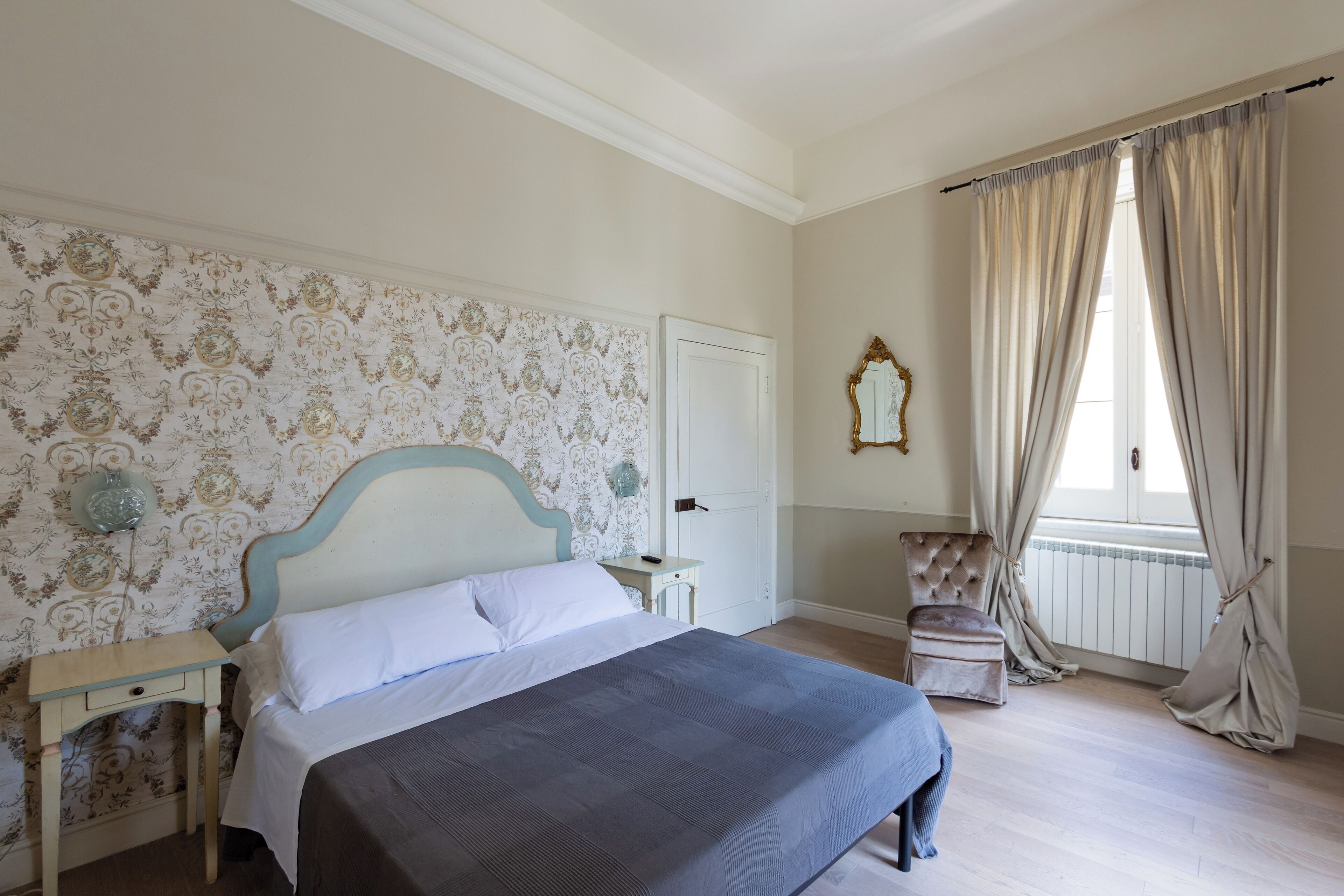 Un dormitorio con cama, mesita de noche, silla y ventana con cortinas.