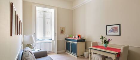 A living room with a sofa, a dining table with a red tablecloth, and a kitchen area with cabinets and a window.