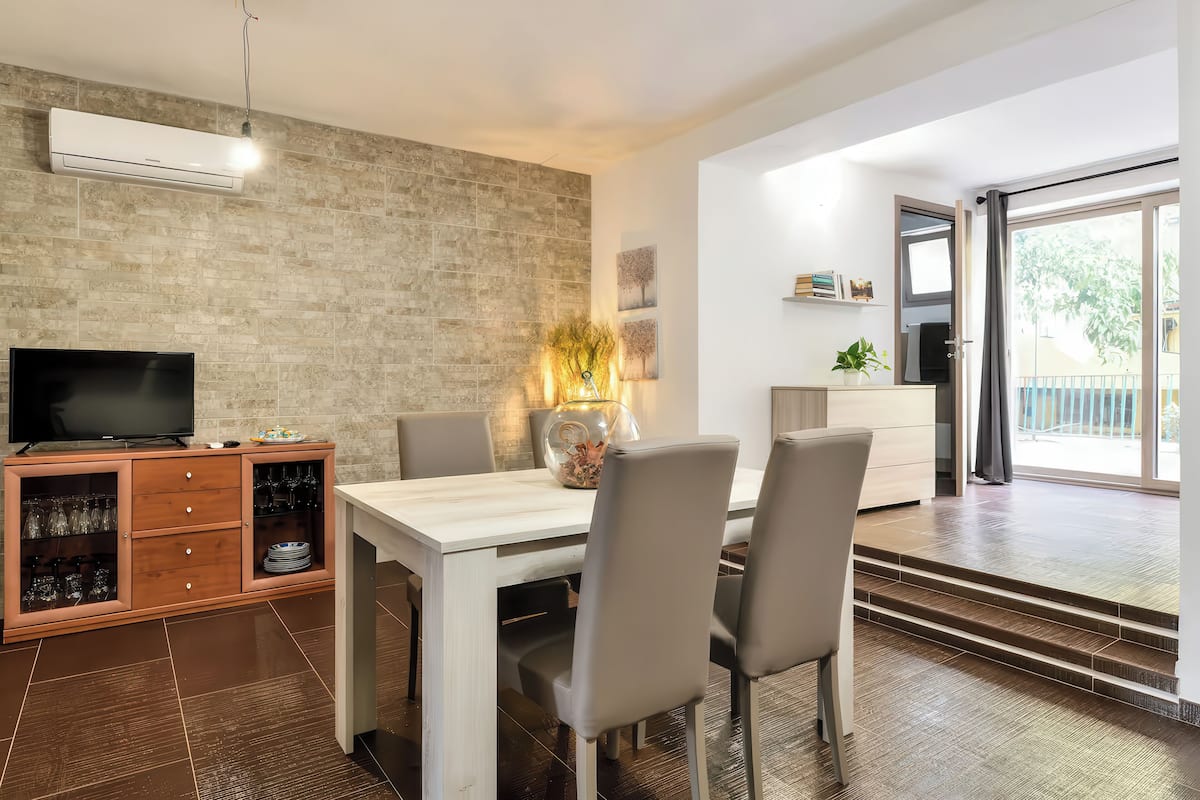 Una sala de estar moderna con televisor, mesa de comedor, sillas y una cocina con puerta corredera al exterior.