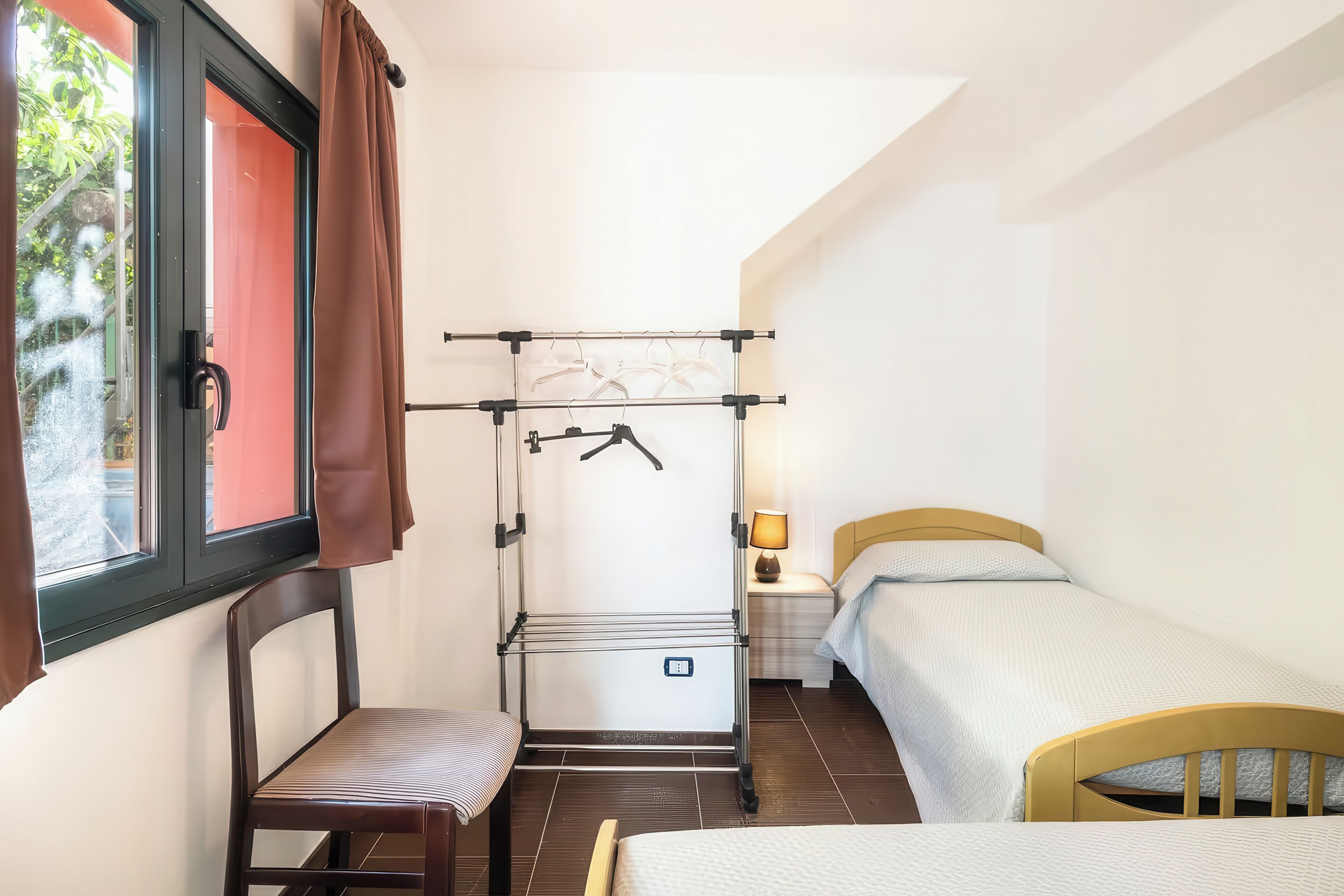 A compact room with a bed, a chair, a small table, a drying rack, and a window with curtains.