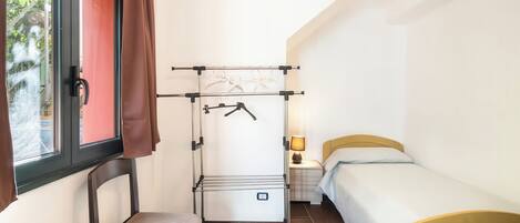 A compact room with a bed, a chair, a small table, a drying rack, and a window with curtains.