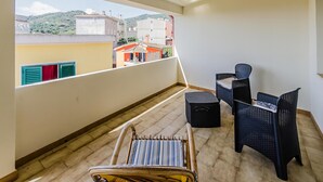 A balcony with a view of buildings and hills, wicker chairs, a small table, and a chair with a striped cushion. - B&B AniMo (Loceri)