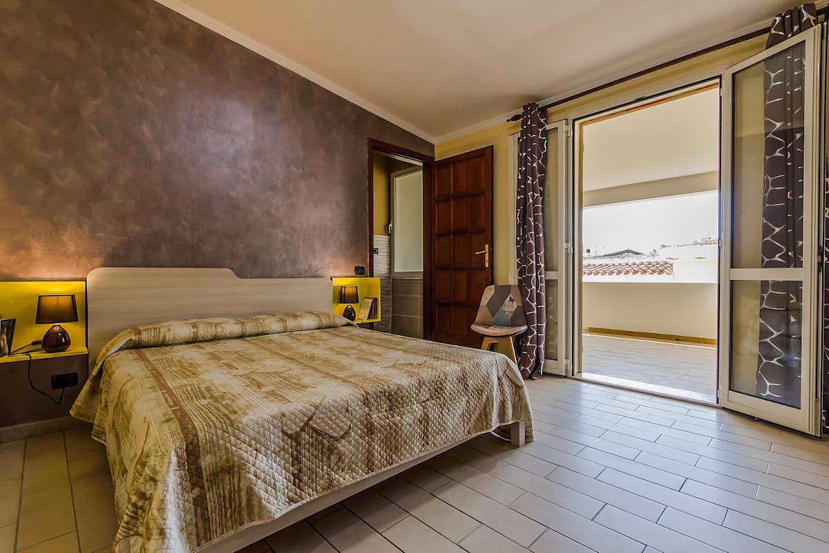 Habitación de hotel con una cama grande, una mesita de noche con lámpara, una silla y un ventanal corredero que da a un balcón.