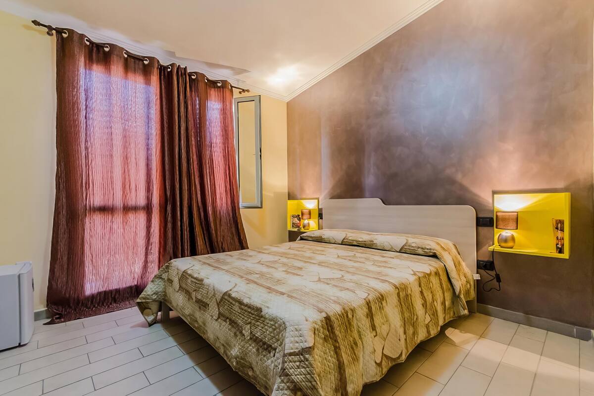 Un dormitorio con una cama grande, cortinas, una mesita de noche con lámpara y un estante de pared con lámpara.