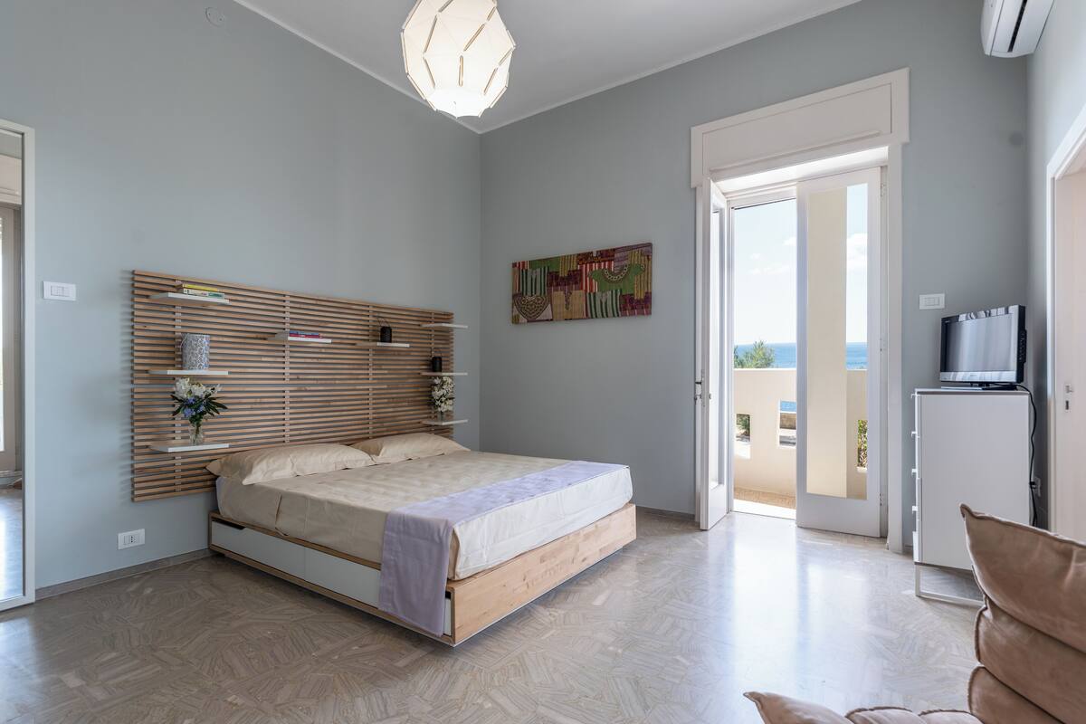 A bedroom with a bed, a wooden headboard, a colorful artwork, a TV, and a balcony with a view.