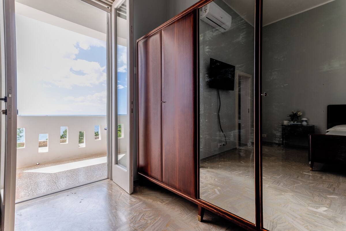 A room with a sliding glass door leading to a balcony, a wardrobe, a bed, and a television mounted on the wall.