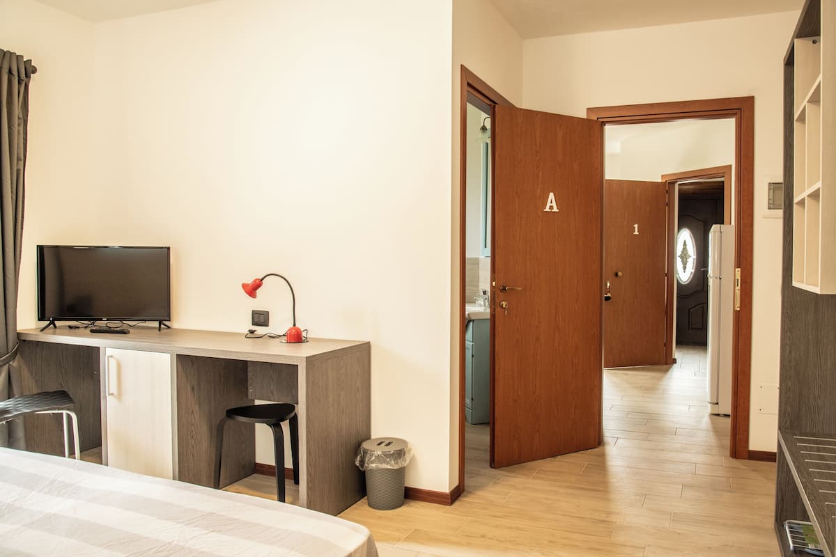 A hotel room with a bed, a desk with a TV, a small chair, a trash bin, and a wooden door with a number.