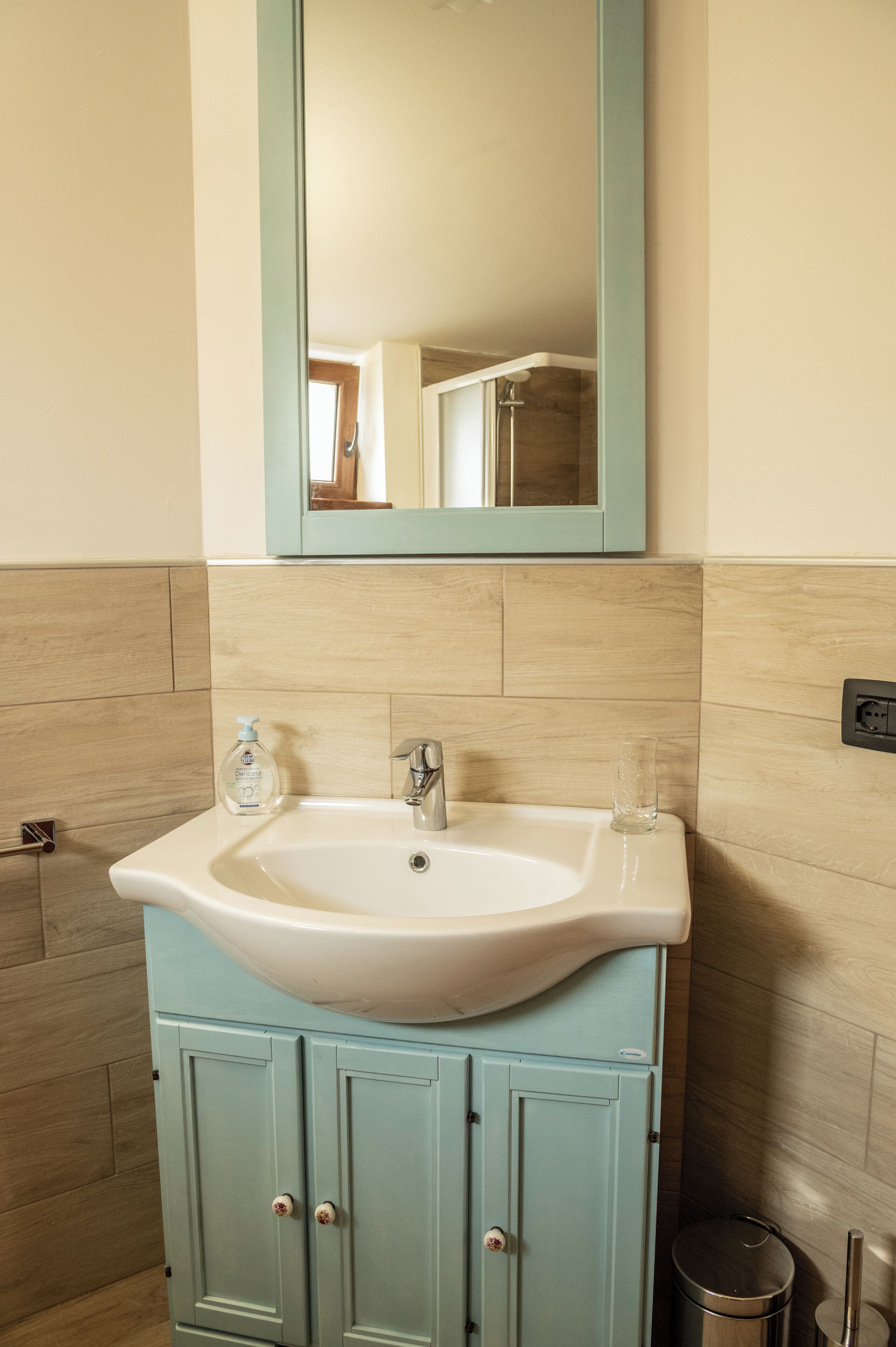 A bathroom with a white sink, light blue cabinets, a mirror above the sink, and a small trash bin to the right.